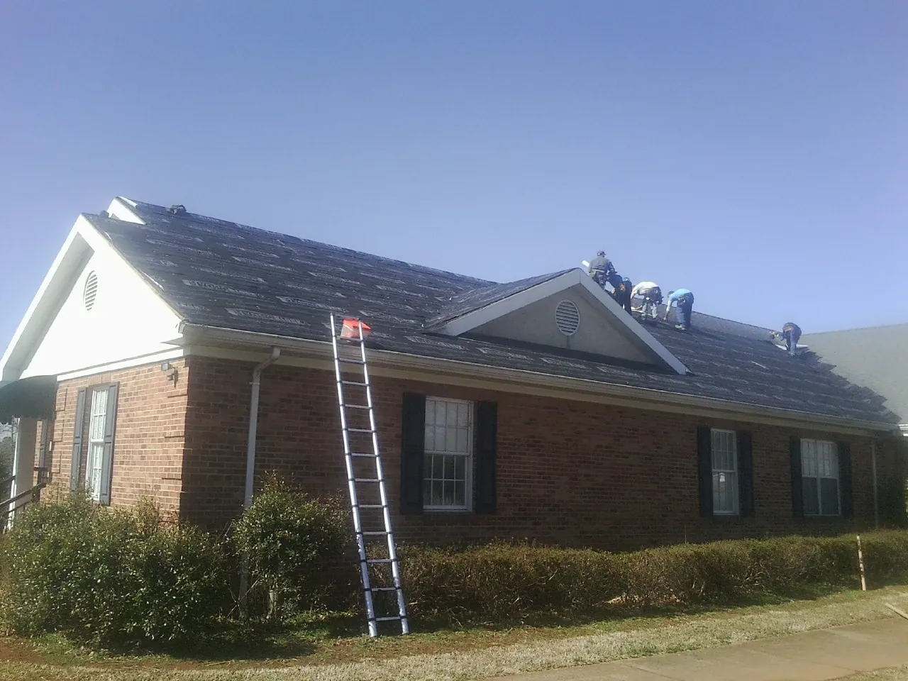 Crew tearing shingles off a brick ranch roof with a ladder set — Metal Roof Repair work by our crew in Dora