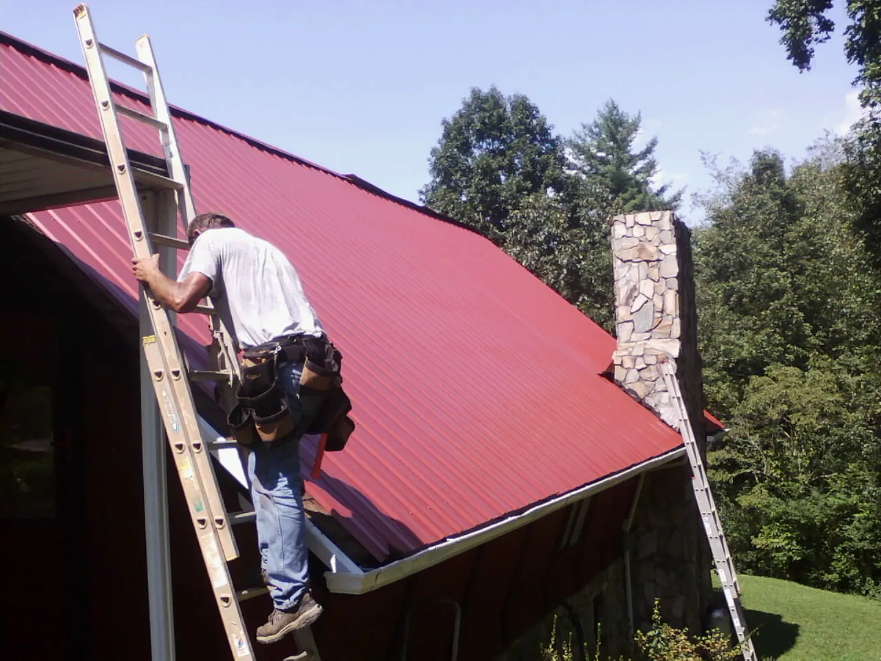 Roofer on a ladder beside a red metal roof and stone chimney — Soffit & Fascia Repair by Roofing Brothers in Dora